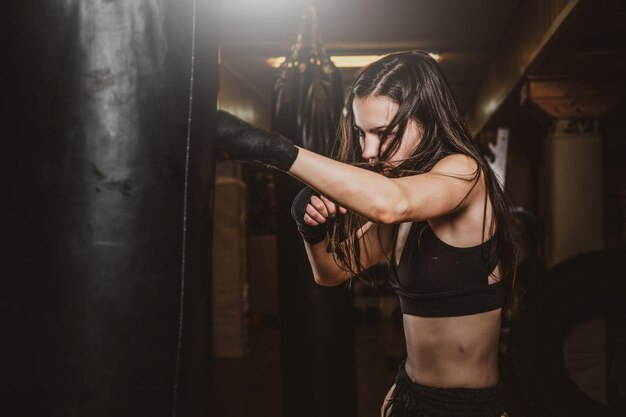 Una mujer flaca enfocada tiene un entrenamiento de boxeo con saco de boxeo en un gimnasio oscuro.