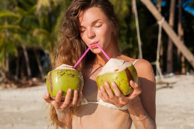 Mujer flaca joven en traje de baño bikini blanco sosteniendo cocos, sonriendo, tomando el sol en la playa tropical.