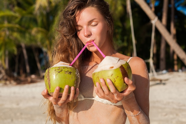 Mujer flaca joven en traje de baño bikini blanco sosteniendo cocos, sonriendo, tomando el sol en la playa tropical.