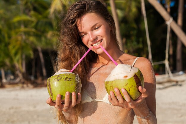Mujer flaca joven en traje de baño bikini blanco sosteniendo cocos, sonriendo, tomando el sol en la playa tropical.