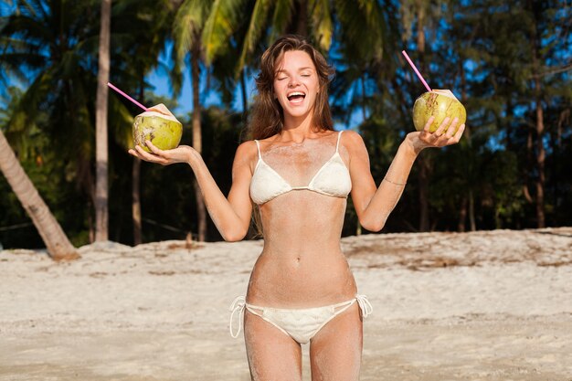 Mujer flaca joven en traje de baño bikini blanco sosteniendo cocos, sonriendo, tomando el sol en la playa tropical.