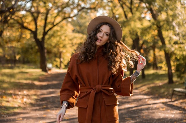 Mujer flaca sonriente elegante atractiva con el pelo rizado caminando en el parque vestida con abrigo marrón cálido, estilo callejero de moda de otoño