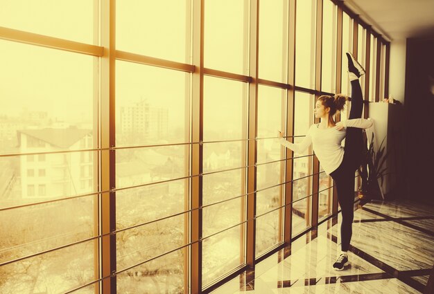 Mujer flexible estirando su pierna izquierda y mirando a través de la ventana