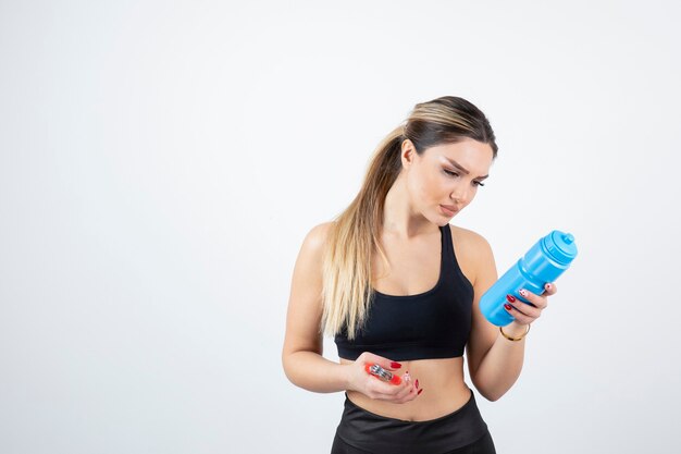 Mujer en forma rubia en top negro de pie y sosteniendo una botella de agua con expansor.