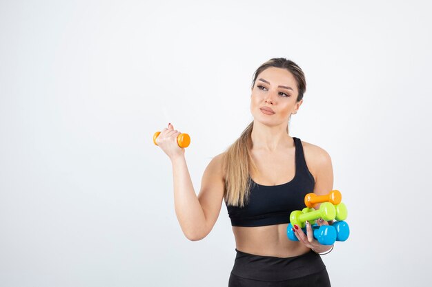 Mujer en forma rubia en top negro de pie y sosteniendo pesas de colores.