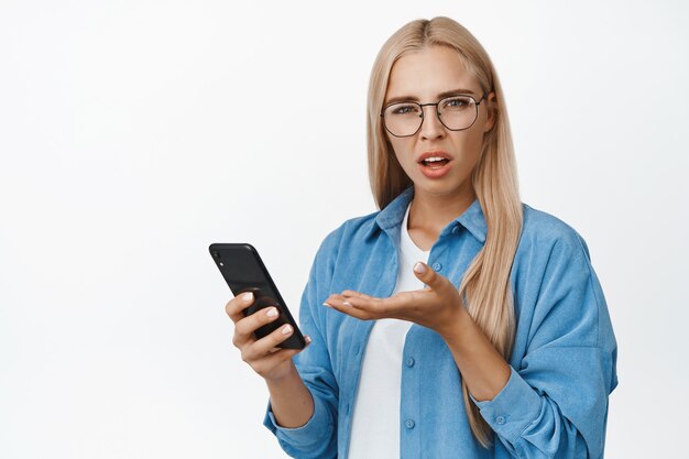 Mujer frustrada con gafas quejándose del teléfono inteligente, sosteniendo el teléfono móvil y mirando desconcertado en blanco.