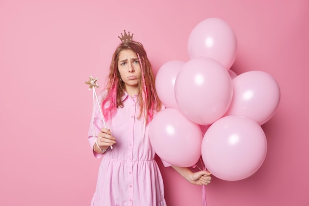 Mujer frustrada infeliz viene en poses de fiesta con globos inflados y varita mágica usa corona de princesa pequeña y poses de vestido sobre fondo rosa Concepto de vacaciones y celebración de personas