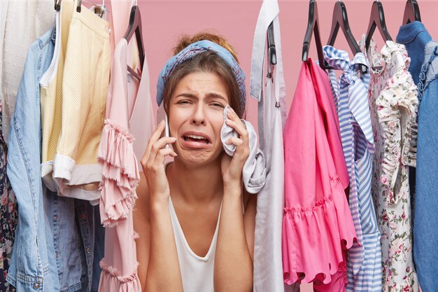 Mujer frustrada de pie cerca del estante lleno de ropa, conversando con su amiga por teléfono inteligente, quejándose de que no tiene nada que ponerse, con una expresión de horror. Ropa, concepto de moda