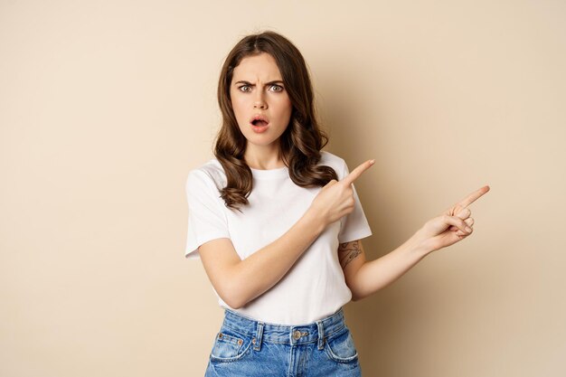 Mujer frustrada que parece insultada señalando con el dedo a la derecha y jadeando sorprendida de pie en camiseta sobre...