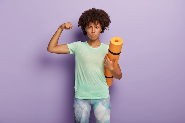 Mujer fuerte y poderosa que levanta el brazo, muestra los bíceps, sostiene la colchoneta de fitness para entrenar en el gimnasio, vestida con ropa deportiva