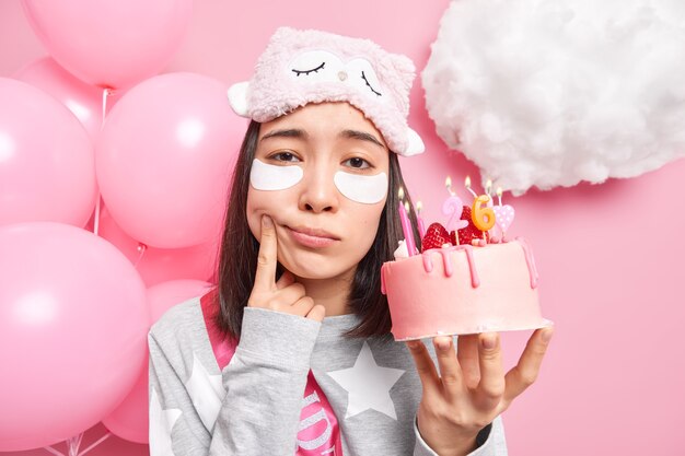 La mujer fuerza la sonrisa se siente molesta por envejecer tiene un delicioso pastel de fresa celebra el cumpleaños solo vestida con ropa de dormir