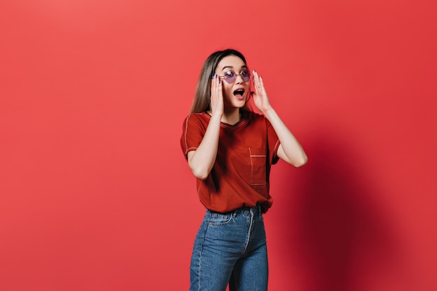 Mujer con gafas de color lila y camiseta roja posando emocionalmente en la pared aislada