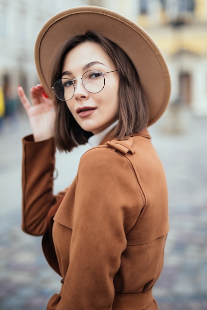 Foto gratuita mujer con gafas modernas y sombrero de moda y abrigo marrón está posando en el centro de la ciudad