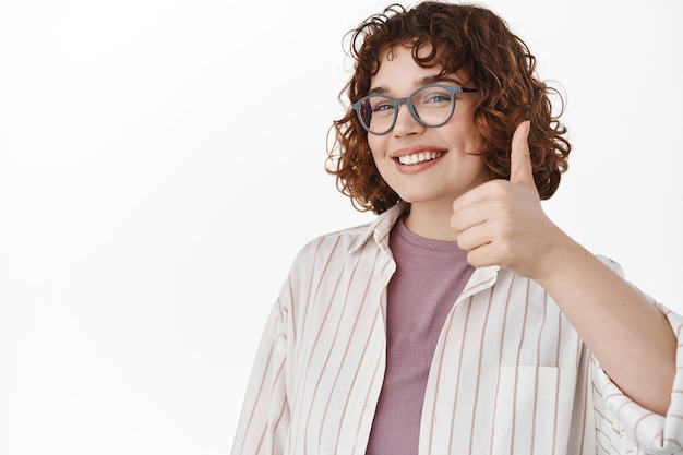 Foto gratuita mujer con gafas mostrando el pulgar hacia arriba en apoyo, aprobar y estar de acuerdo, asentir satisfecha, como algo, elogiar la gran elección en blanco.