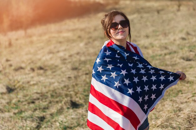 Mujer en gafas de sol envuelto en bandera americana mirando a cámara