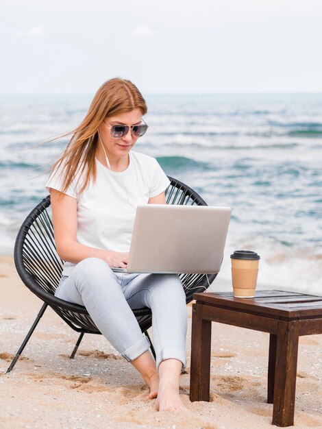 Mujer con gafas de sol en silla de playa trabajando en la computadora portátil