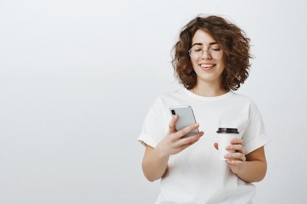 Mujer con gafas sonriendo al mensaje de texto del teléfono móvil, tomando café en el descanso de la oficina