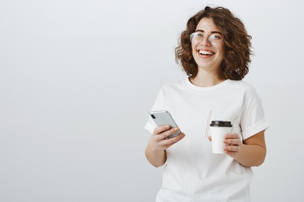 Mujer con gafas sonriendo al mensaje de texto del teléfono móvil, tomando café en el descanso de la oficina