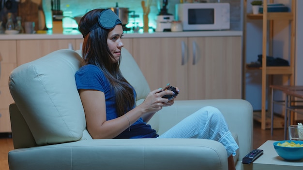 Mujer gamer jugando videojuegos en la consola con controlador y joysticks sentado en el sofá frente al televisor. Persona determinada emocionada que se relaja jugando con el controlador inalámbrico divirtiéndose ganando