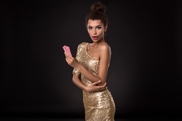 Mujer ganadora: mujer joven con un elegante vestido dorado que sostiene dos cartas, una combinación de cartas de póquer de ases. Foto de estudio sobre fondo negro. emociones