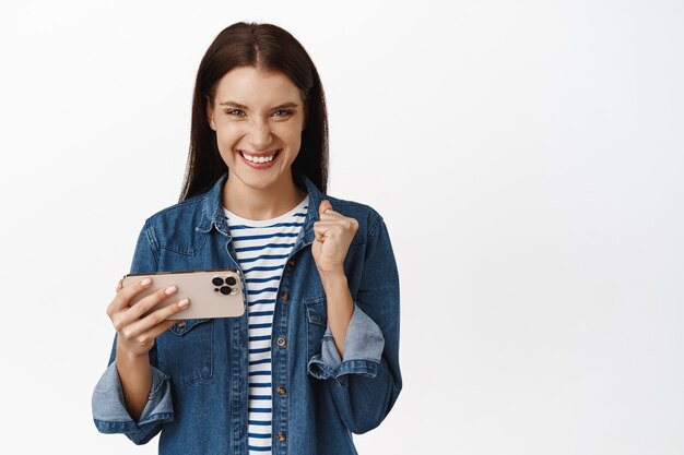 Mujer ganando en videojuegos móviles, sosteniendo el teléfono inteligente en posición horizontal y luciendo complacida, viendo algo en el teléfono en blanco