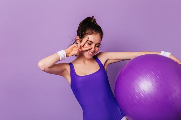Mujer gimnasta delgada sostiene fitball y muestra el signo de la paz en la pared púrpura