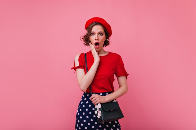 Mujer glamorosa delgada con peinado ondulado que expresa emociones conmocionadas. Fotografía interior de modelo femenino francés en boina.