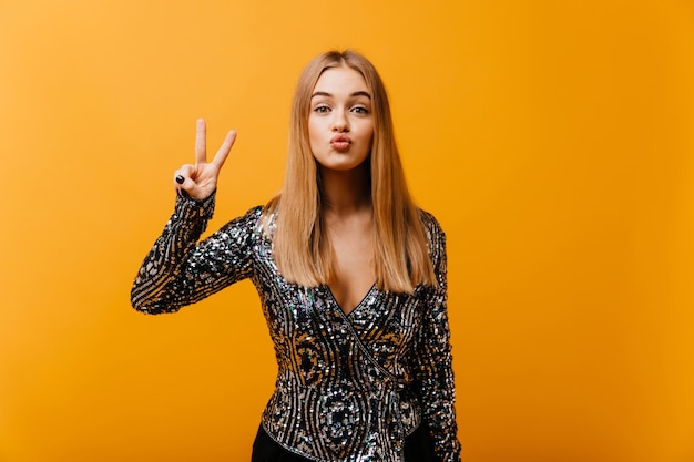 Mujer glamorosa divertida posando en naranja con el signo de la paz. Retrato interior de una encantadora mujer ciega con chaqueta brillante.