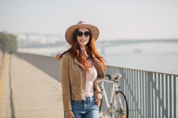 Mujer glamorosa en gafas de sol negras posando en puente con bicicleta en la pared