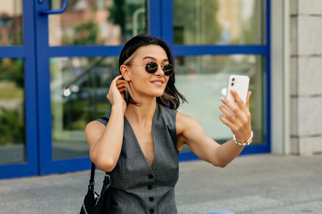 glamorosa en chaleco gris y gafas de sol oscuras está haciendo en teléfono inteligente | Foto Gratis