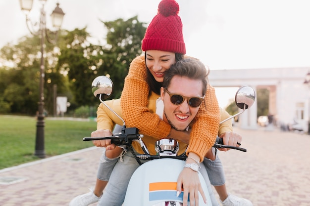 Foto gratuita mujer glamorosa en lindo gorro de punto mirando a otro lado con sonrisa, montando en scooter con novio