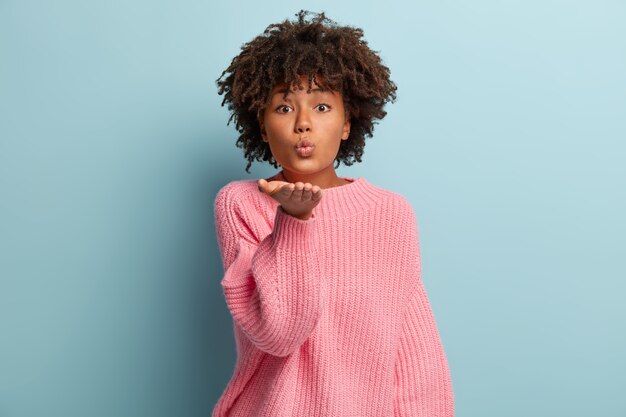 Mujer glamorosa de piel oscura envía beso al aire, mantiene los labios doblados, tiene corte de pelo afro