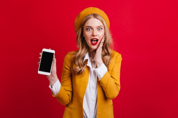 Mujer glamorosa sorprendida que sostiene el teléfono inteligente con pantalla en blanco en la pared roja. Atractiva chica rubia en boina amarilla posando con dispositivo digital