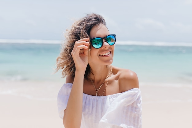 Foto gratuita mujer glamorosa en vestido blanco disfrutando del verano en el resort. retrato de impresionante dama bronceada con gafas de sol de pie cerca del mar.