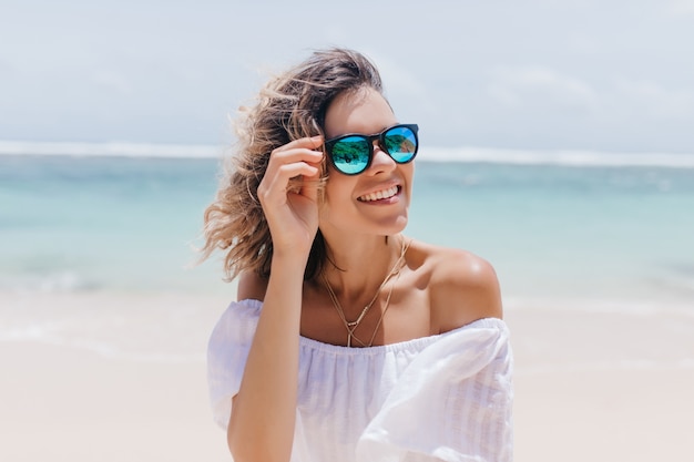 Mujer glamorosa en vestido blanco disfrutando del verano en el resort. Retrato de impresionante dama bronceada con gafas de sol de pie cerca del mar.