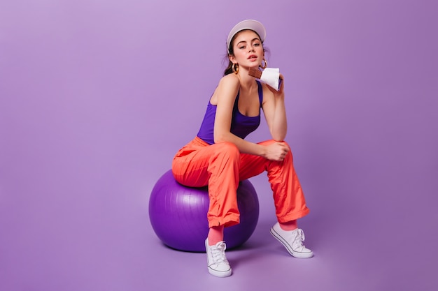 Mujer con gorra y traje deportivo violeta-naranja se sienta en fitball y sostiene chocolate con leche