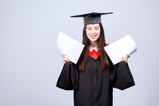 Mujer con gorro de graduación y bata de ceremonia mostrando papeles vacíos. Foto de alta calidad