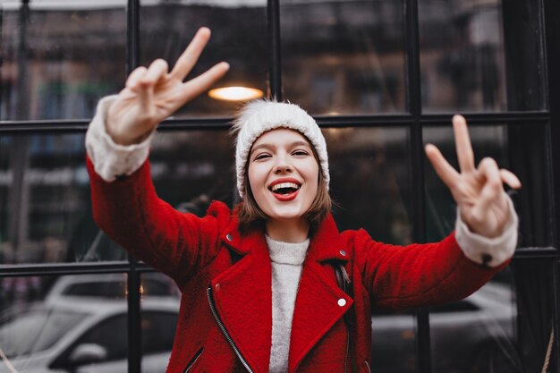 Mujer con gorro de punto y chaqueta roja está sonriendo, mostrando signos de paz y mirando a la cámara contra el fondo de la ventana con marco de madera negro.