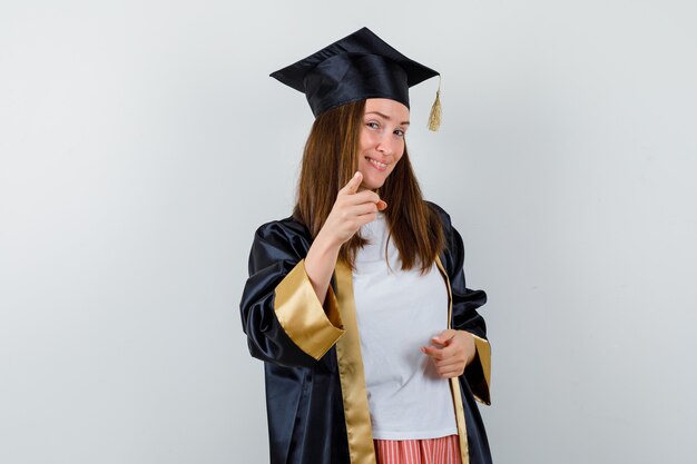 Mujer graduada apuntando a la cámara en ropa casual, uniforme y confiada. vista frontal.