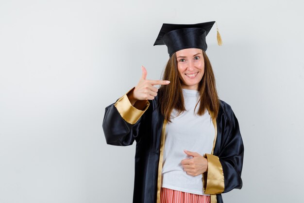 Mujer graduada apuntando a la derecha en ropa casual, uniforme y feliz. vista frontal.