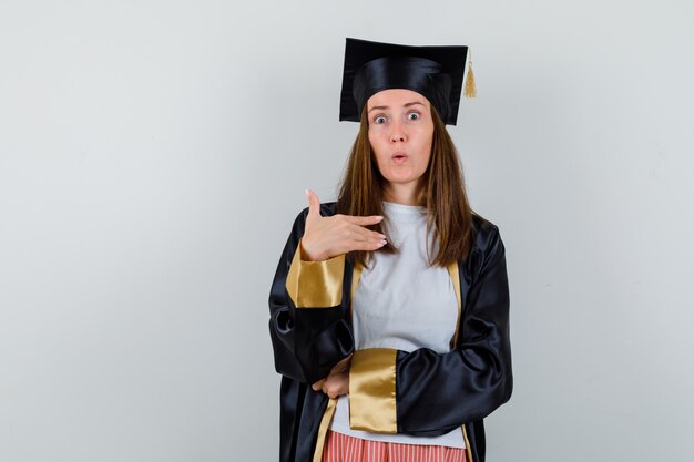 Mujer graduada apuntando a sí misma en ropa casual, uniforme y mirando sorprendida. vista frontal.