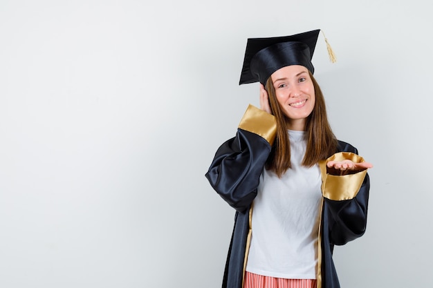 Mujer graduada extendiendo la mano a la cámara, sosteniendo la otra mano en la oreja con ropa casual, uniforme y luciendo esperanzada, vista frontal.