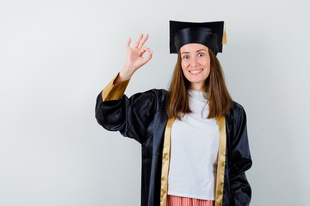 Mujer graduada mostrando gesto ok en ropa casual, uniforme y mirando alegre, vista frontal.