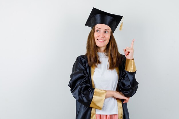 Mujer graduada en ropa casual, uniforme apuntando hacia arriba y mirando esperanzado, vista frontal.