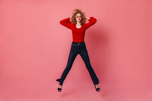 La mujer de gran humor alegre está saltando en el espacio rosa. dama de anteojos, blusa roja y pantalones silbando y tocando su pelo rizado.