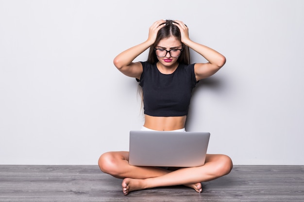 Mujer gritando sorprendida sentada en el suelo con la computadora portátil mientras sostiene su cabeza sobre fondo gris