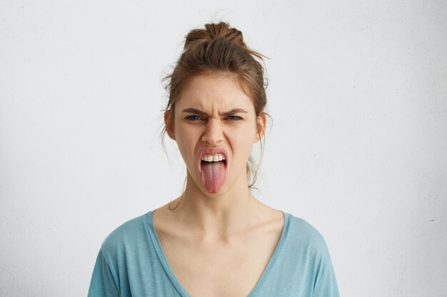 Mujer gruñona con nudo de pelo mostrando su lengua expresando emociones negativas. Mujer furiosa mostrando su disgusto mientras pelea