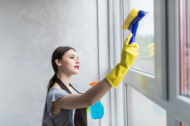 Mujer con guantes de protección está sonriendo y limpiando el polvo con un spray y un plumero mientras limpia su casa, primer plano