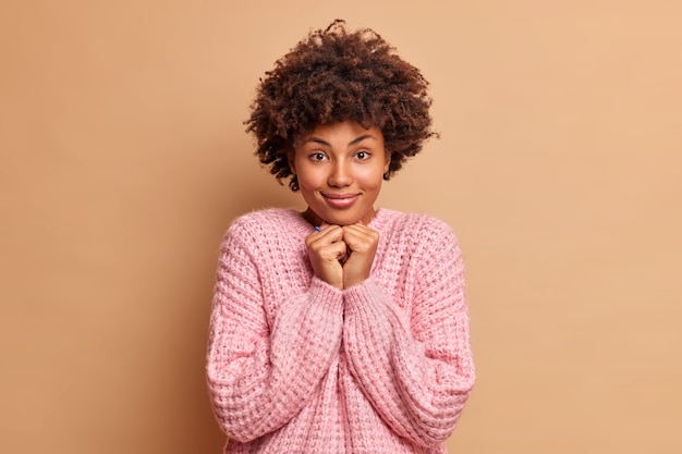 Una mujer guapa con cabello rizado mantiene las manos debajo de la barbilla mira directamente al frente escucha algo atentamente viste un suéter de punto casual aislado sobre una pared marrón admira algo maravilloso