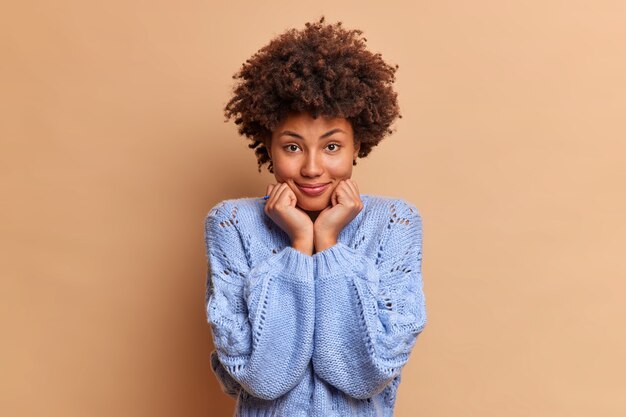 Una mujer guapa con cabello rizado natural mantiene las manos debajo de la barbilla vestida con un jersey azul mira directamente al frente se encuentra confiado y hermoso se encuentra contra la pared marrón